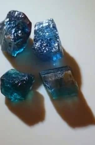 Four rough blue and teal tourmaline crystals turning under light on a white surface.
