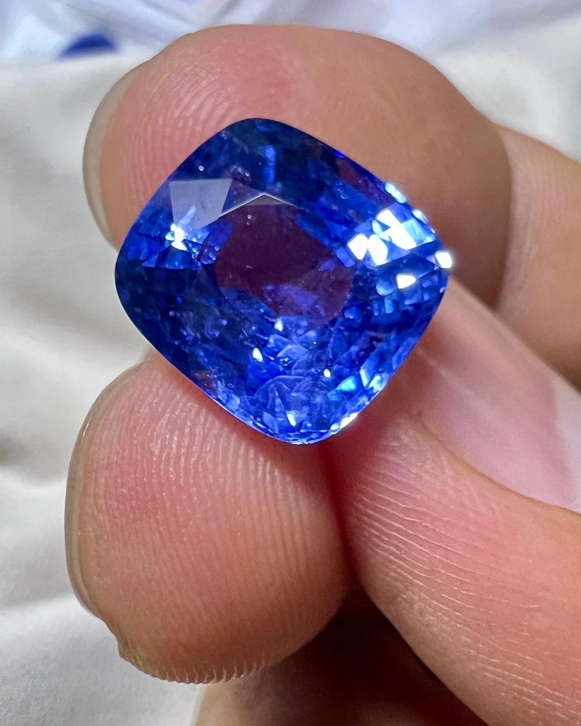 A large cushion-cut blue sapphire held between finger and thumb, showing strong saturation and bright facet reflections.