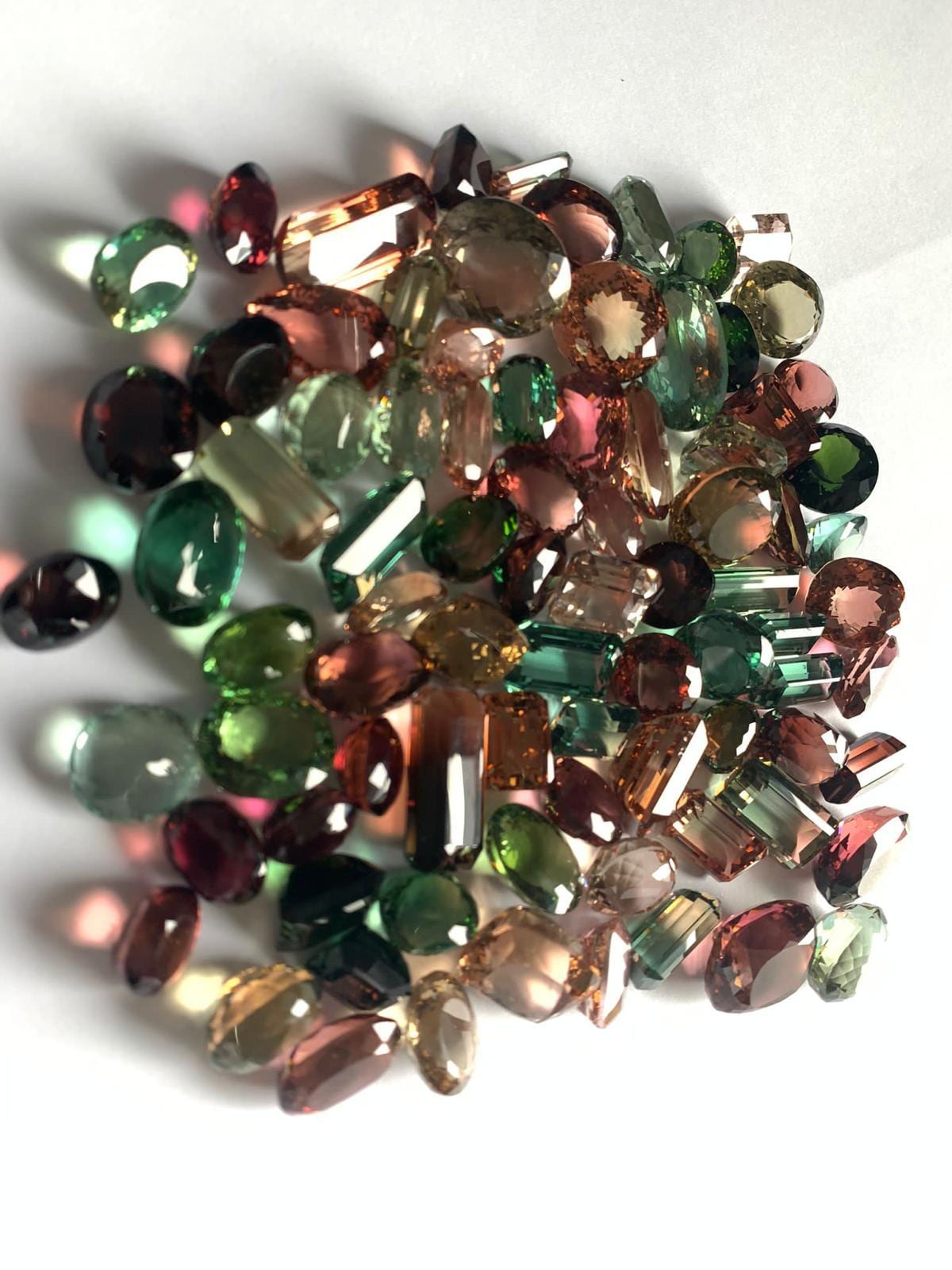 Loose faceted tourmalines scattered on white — greens, pinks, reds and warm peach tones across mixed shapes and sizes.