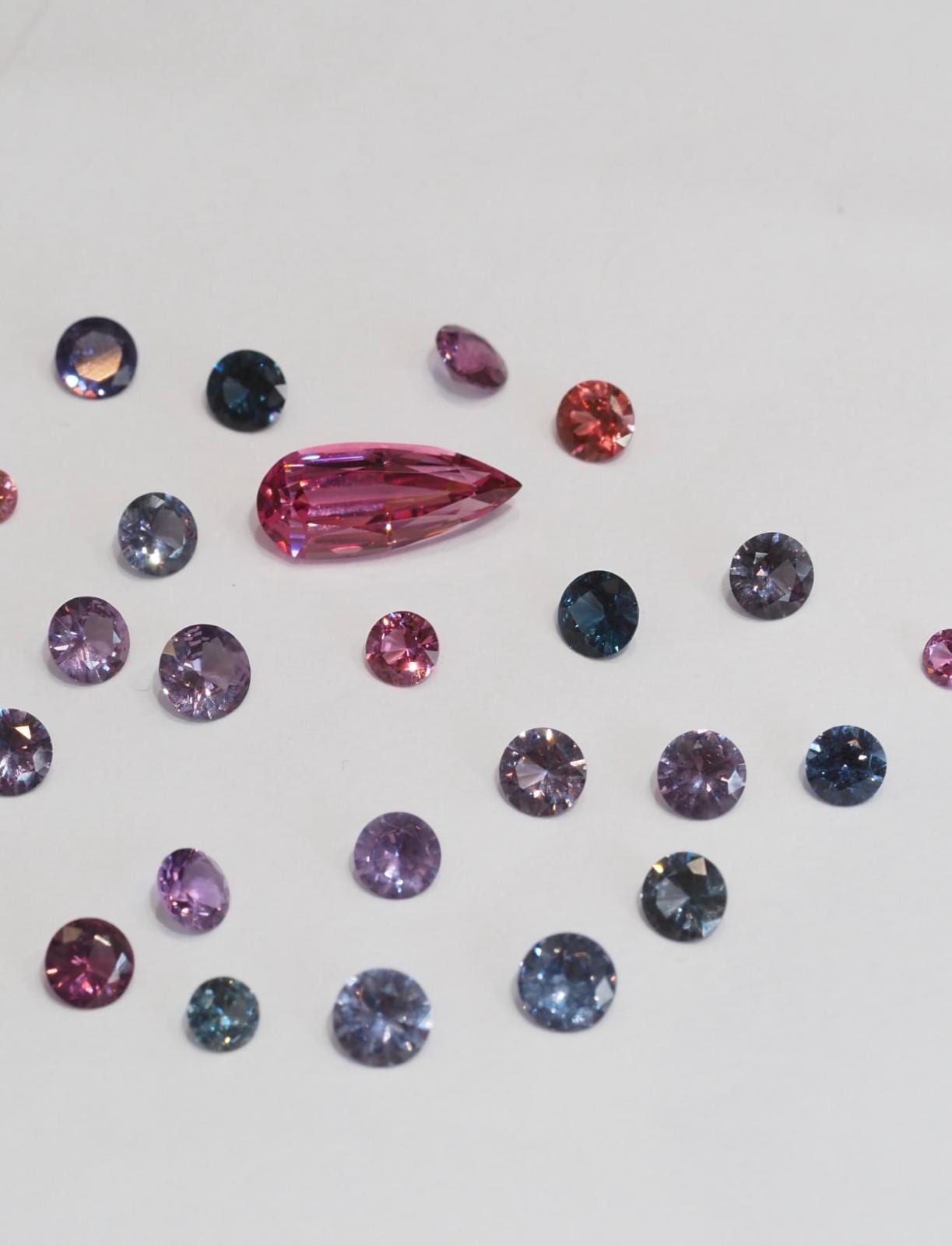 Spinels spaced on a white ground in lavender, steel grey, mauve and pink, mostly round brilliants with a pink pear-cut stone at the centre.
