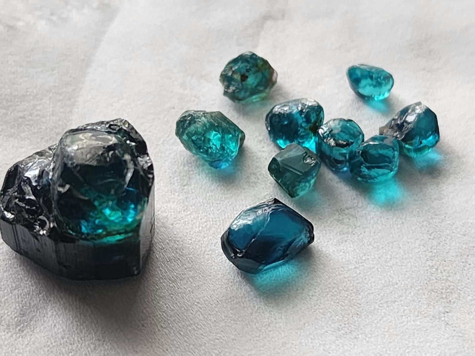 A large rough tourmaline crystal with a dark termination and vivid blue-green core, beside nine smaller rough pieces in blue and teal on a white cloth.