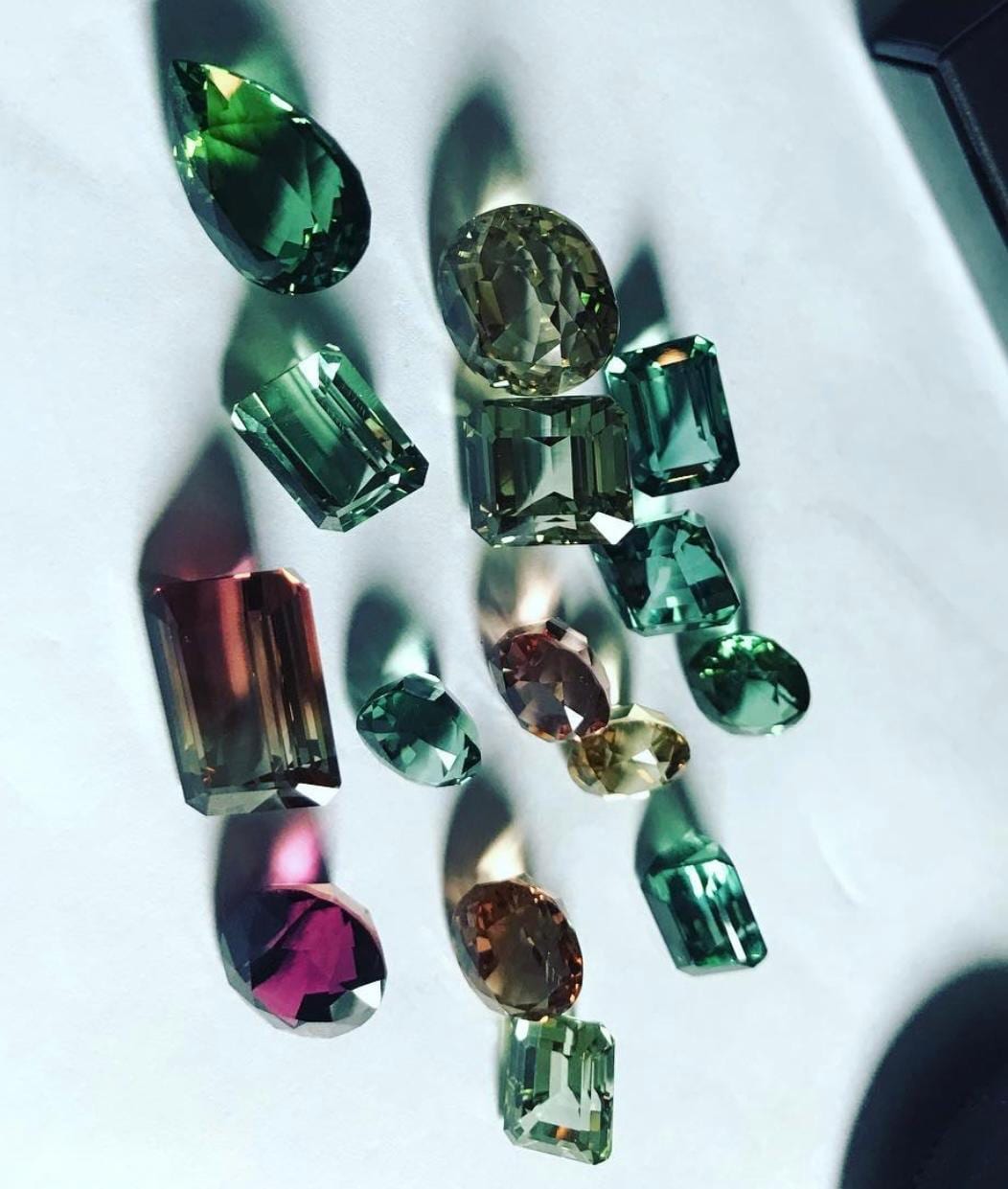 A suite of green and teal tourmalines in direct sunlight — pear, oval and emerald cuts casting coloured shadows, with one red-brown bi-colour stone.